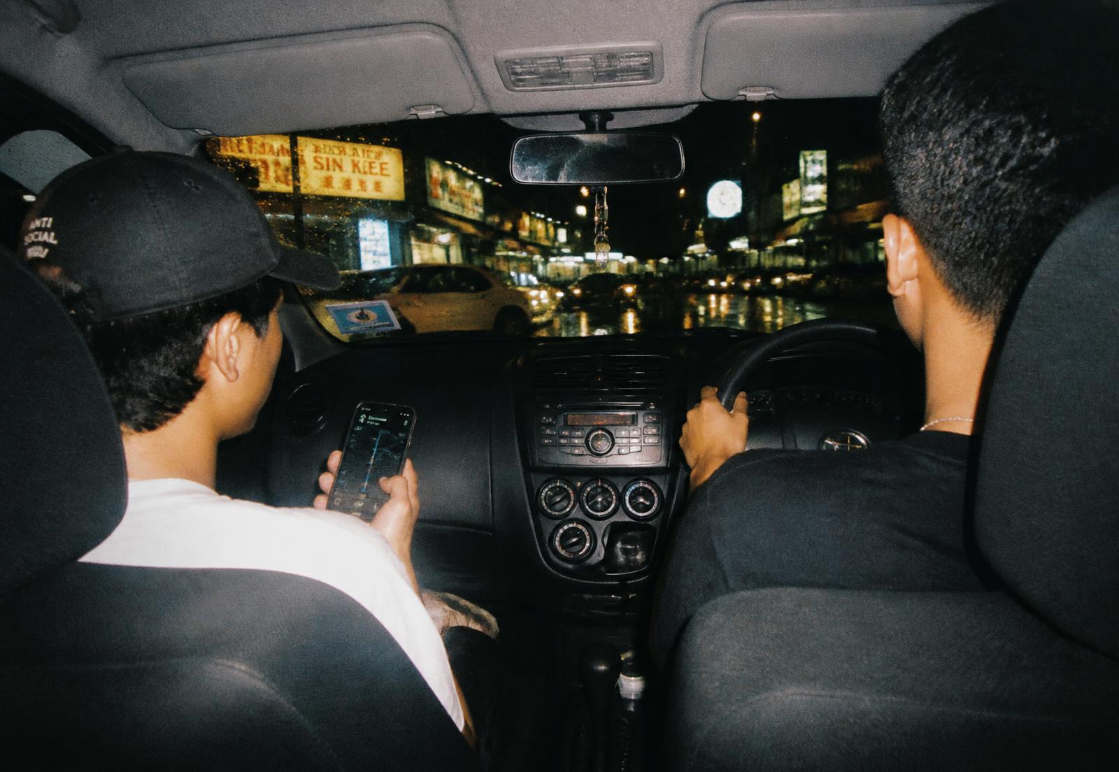 Two friends driving around Kuala Lumpur at night, one holding a phone with a map open
