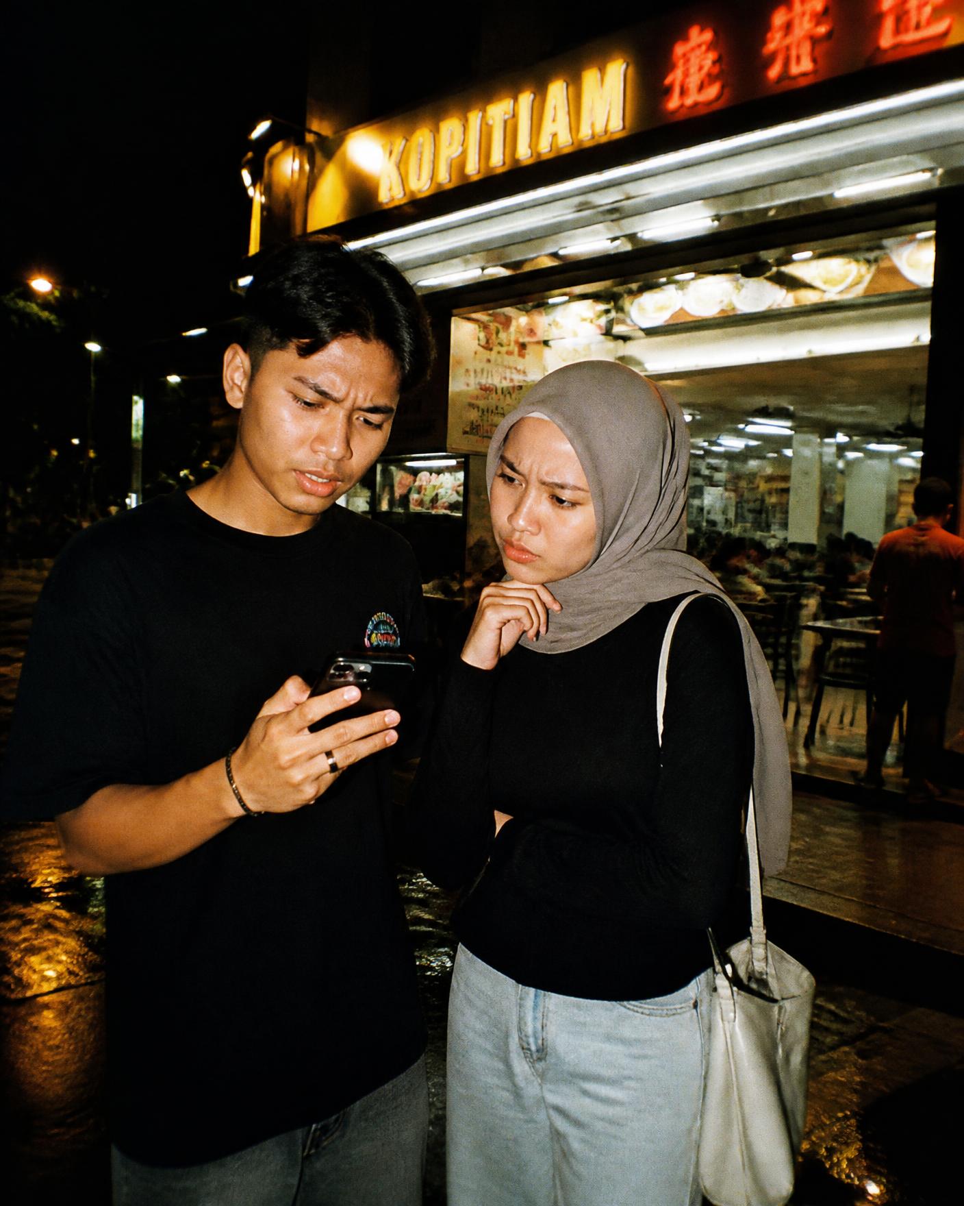 A couple standing outside a kopitiam at night, staring at one phone, undecided